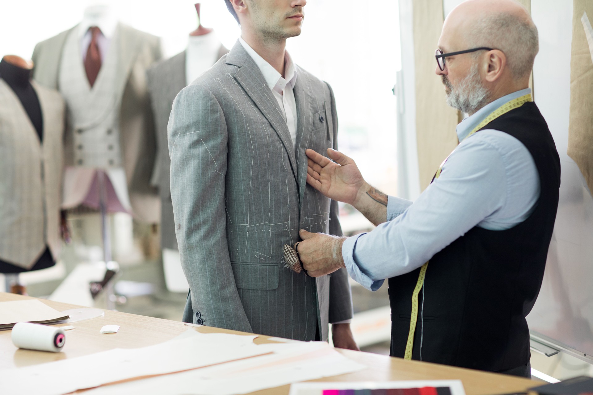 Confident bald senior male tailor in glasses checking suit jacket in client and offering ideas to him for custom-made suit in workshop